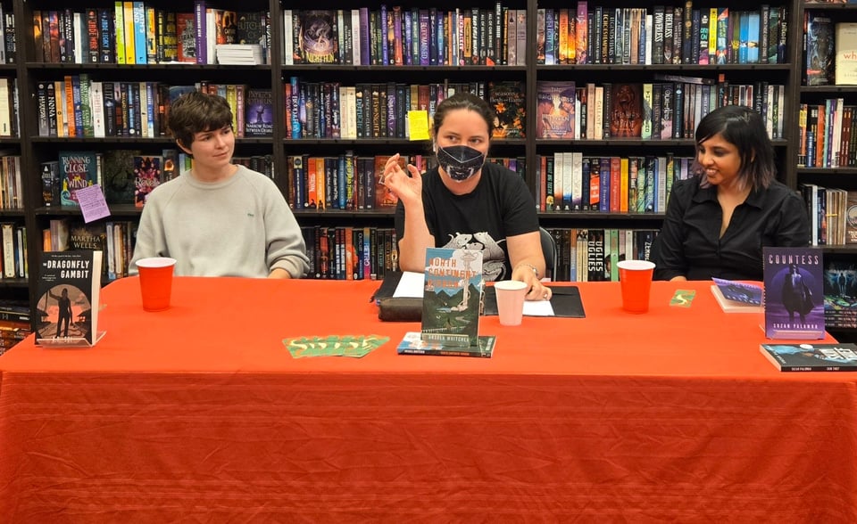 A.D. Sui in a trim sweatshirt, me in a mask, and Suzan Palumbo in her favorite goth black, sitting at a table behind our books at the Bakka Phoenix bookstore, with even more books in the background