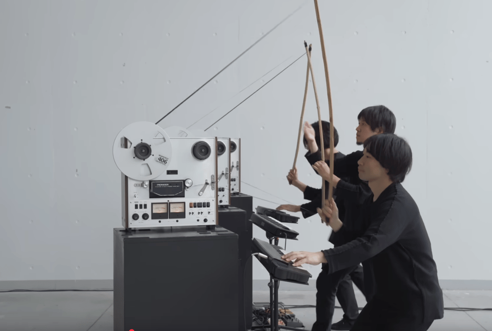 Three musicians dressed in black perform with a reel-to-reel tape machine and electronic instruments. Each holds a long wooden bow, drawing it across tall metal rods attached to the tape decks, creating an experimental and futuristic performance setup in a minimalist white room.
