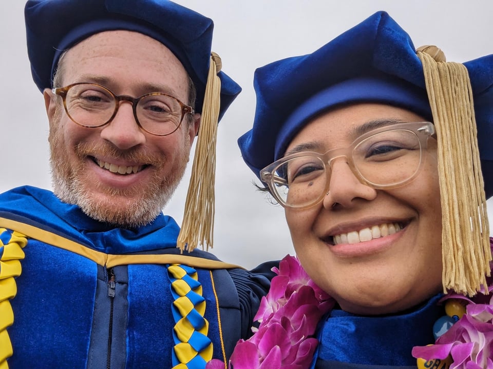 Two smiling humans wearing glasses in the University of California, Santa Cruz commencement regalia in their school colors of blue and gold.