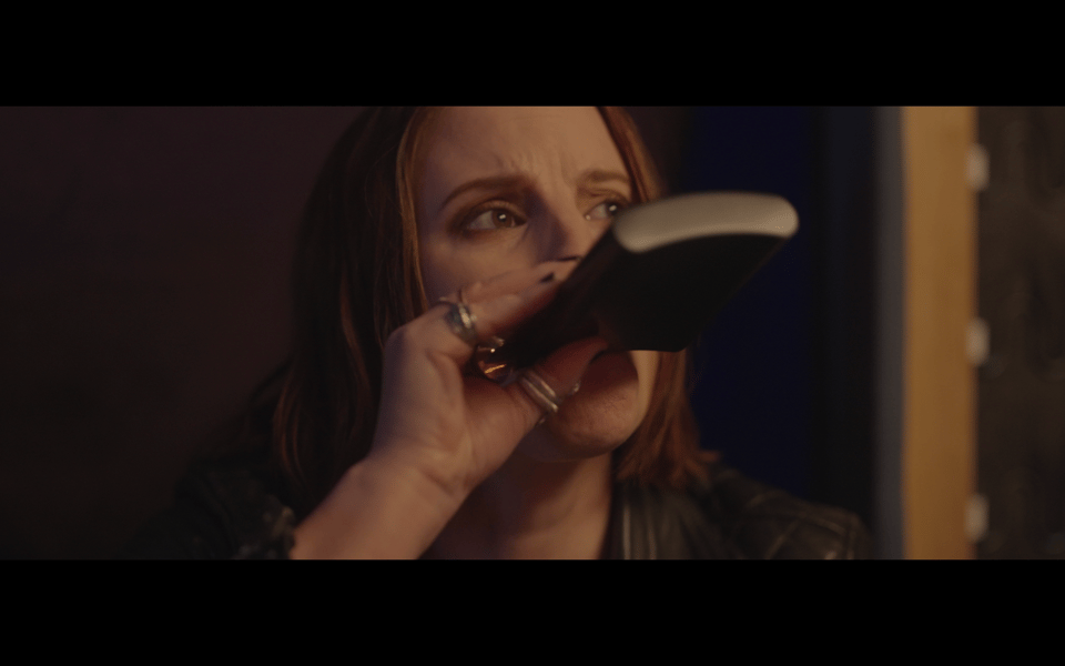 A screenshot of a frame of film. A woman with a forlorn look in her eyes, tips back a flask to drink