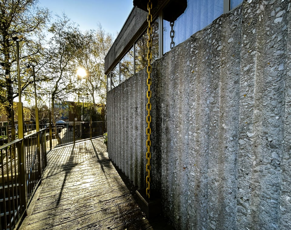 Low winter sun hitting the damp decking and creating shadows on the ribbed concrete wall. In the middle, a mustard yellow rain chain disappears through a gap in the decking.