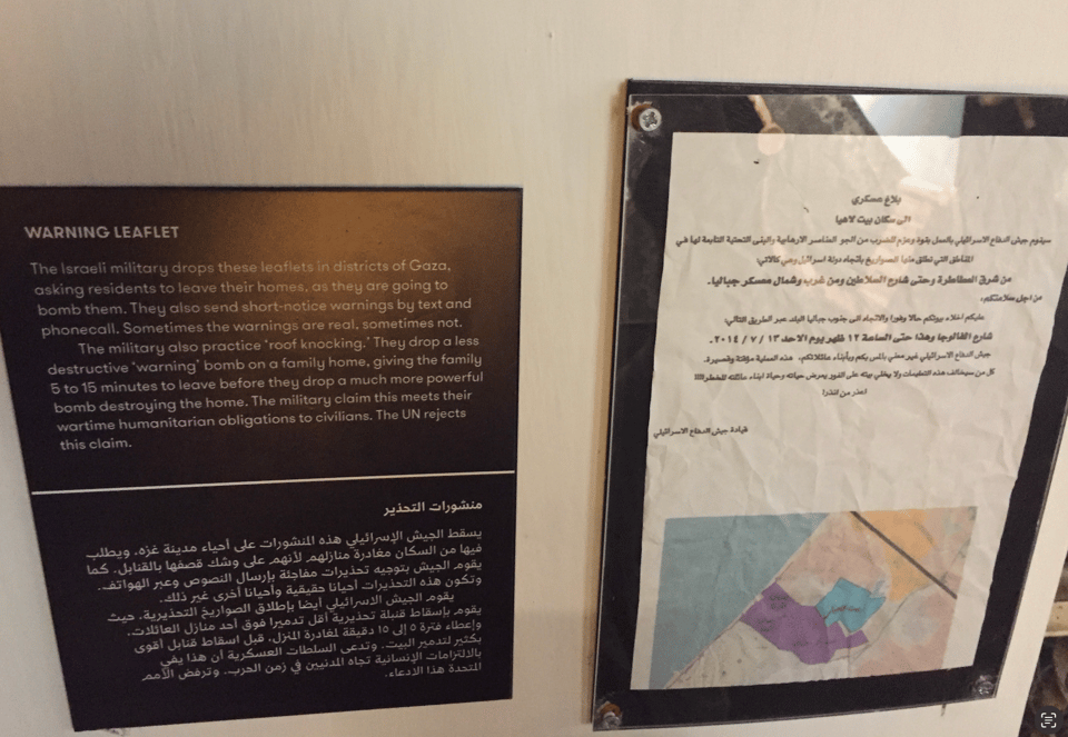 Photograph of an informational sign in English and in Arabic with the heading 'WARNING LEAFLET' next to a framed piece of paper that shows Arabic writing at the top, and a colorful map-like image at the bottom of the page.  (The English version of the text in the informational sign is shown in the caption of this image.)