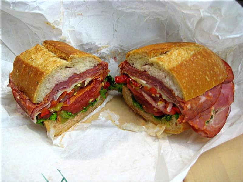 Photo of a hero sandwich cut in half, made with cold cut meats, tomato, lettuce, and other toppings, on a piece of white butcher’s paper. Source: Wikimedia Commons.