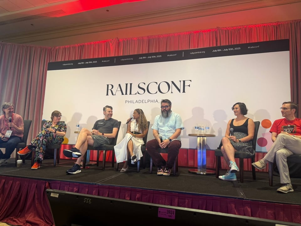 I’m seated on stage with six other panelists at RailsConf 2025 in Philadelphia. The backdrop behind us has the RailsConf branding and event dates (July 8th–10th, 2025). I’m wearing a sleeveless white dress with black piping and a wide black belt, speaking into a microphone with a smile, mid-discussion.