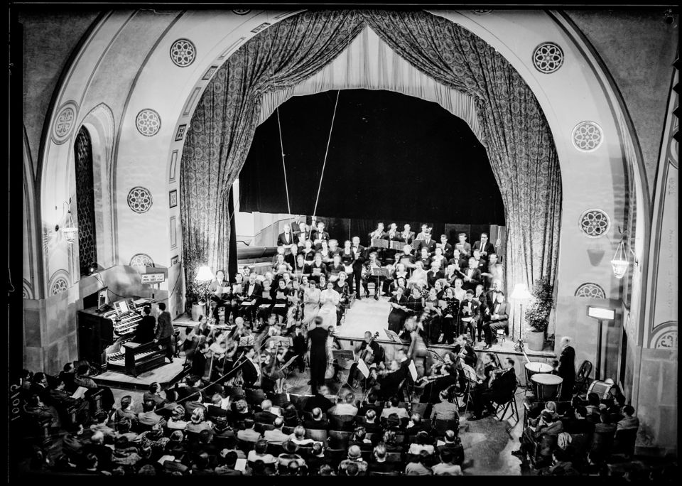 A filled concert hall with an orchestra on a grand stage