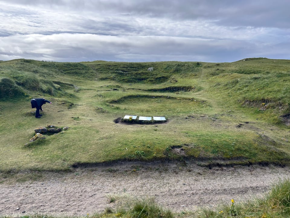In a hilly, grassy landscape by a sandy trail, several circular depressions in the ground stand, lined with drystone wall. Three information panels gives information on Cladh Hallan.