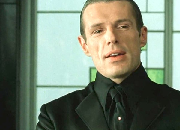 The "Merovingian" villain from the movie "The Matrix Reloaded". He is a short-haired white man dressed in a black suit, with a black shirt and a black tie tied in a very complex knot