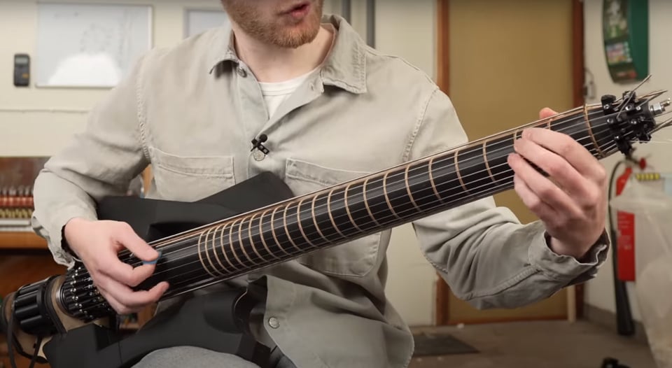 A screenshot of a video showing a man in a beige shirt playing an unusual guitar. It has a black solid body and a black neck that is cyclindrical. There are strings going lengthwise up all sides of the cylinder, and the cylinder has frets running around the width of the tube.