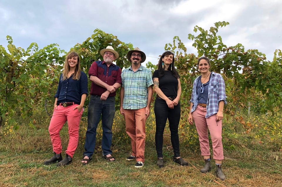 Five people -- three women and two men -- stand next to each other outdoors in front of vegetation.