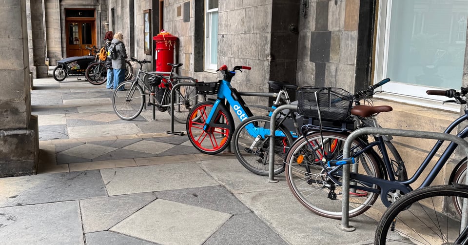 Cycle racks outside an old, stone building - Edinburgh's City Chambers. The cycle racks are nearly at capacity, and standing out among the cycles is a bright blue and red hire bike from ebike provider Dott waiting to be demoed.