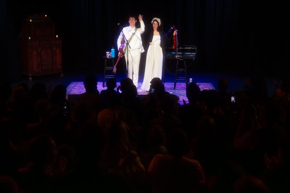 Micah and Marnina, dressed in white wedding outfits, hold hands up, smiling widely at curtain call on a proscenium stage lit with purple. In the foreground is the silhouette of a whole audience on its feet.