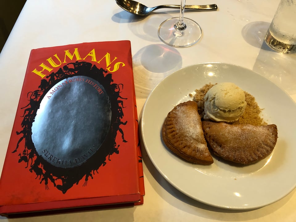 A plate with two semi-circular pies next to a scoop of ice cream on a bed of crumbs on a table with white tablecloth. Two glasses and a spoon at the edge of the photo. A copy of HUMANS: A Monstrous History is to the left of the plate.