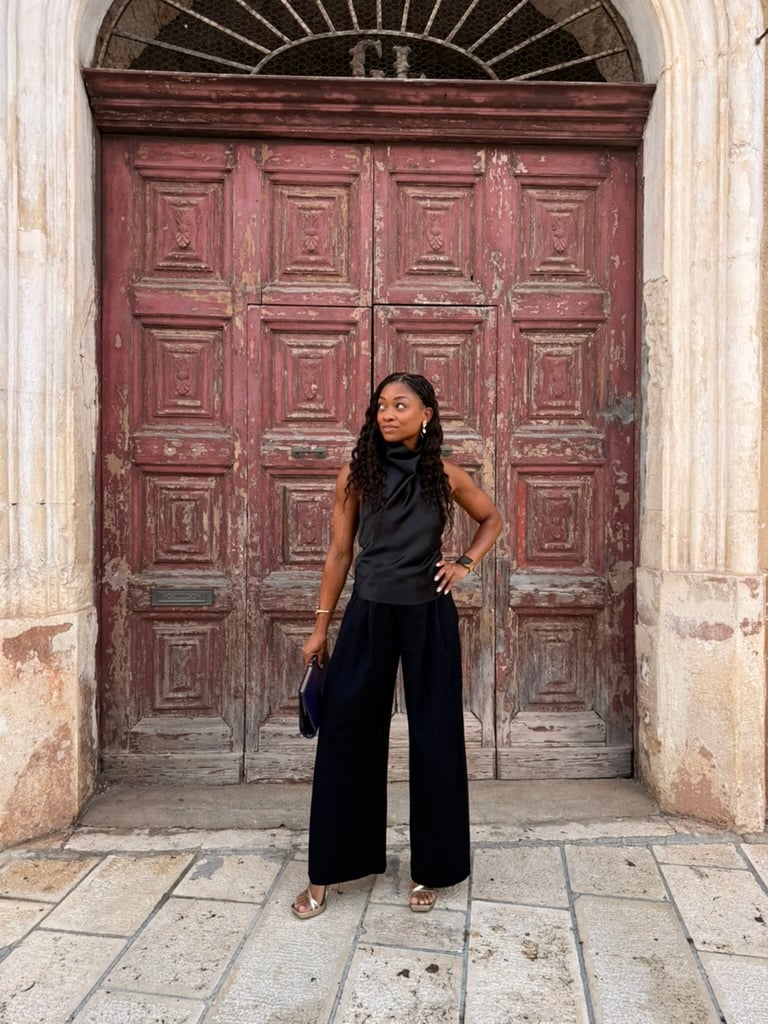 Nadia poses in an all-black outfit in front of a large, weathered red wooden doorway, looking to the side with one hand on her hip.