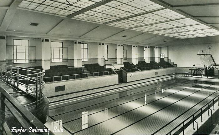 Exeter Swimming Bath in the 1940s. It's a long pool under an equally long skylight set into a concrete beam roof. Ranked seats sit high above the poolside, so spectators are not splashed. At the far end are the diving platforms. In the foreground is the only curve in the place, on a balcony overlooking the shallow end.