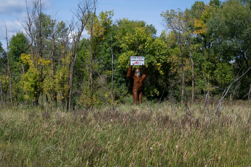 A metal statue of bigfoot, holding a sign for a metal fabrication business. Surrounded by tall grass and trees.