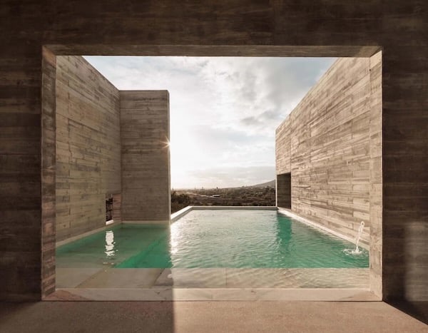 A través de una apertura rectangular en homigón vemos una piscina de aguas azules en forma de L hacia la izquierda. Al lazo izquierdo hay un muro más corto y con una apertura menor y al derecho un muro más largo con una apertura mayor. La pared izquierda oculta un destello de sol y al fondo tras la piscina se intuye el paisaje y el cielo. Todas las paredes son de hormigón.