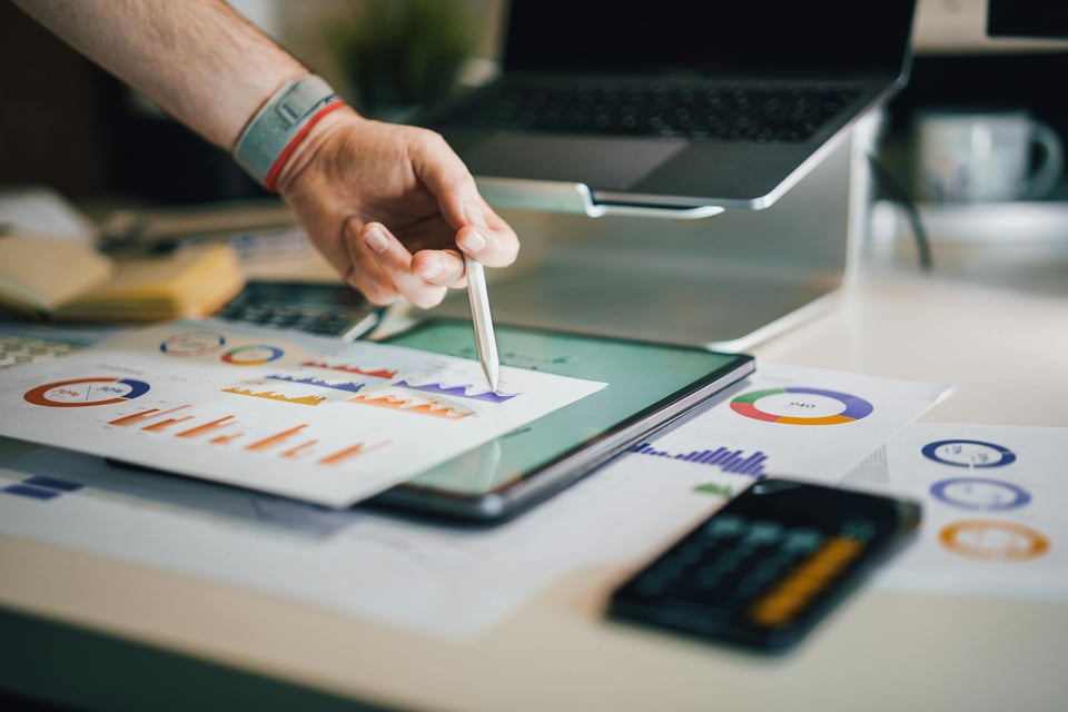 An image of colorful graphs and charts, plus an iPad and a cell phone set on calculator mode, all implying work and productivity.
