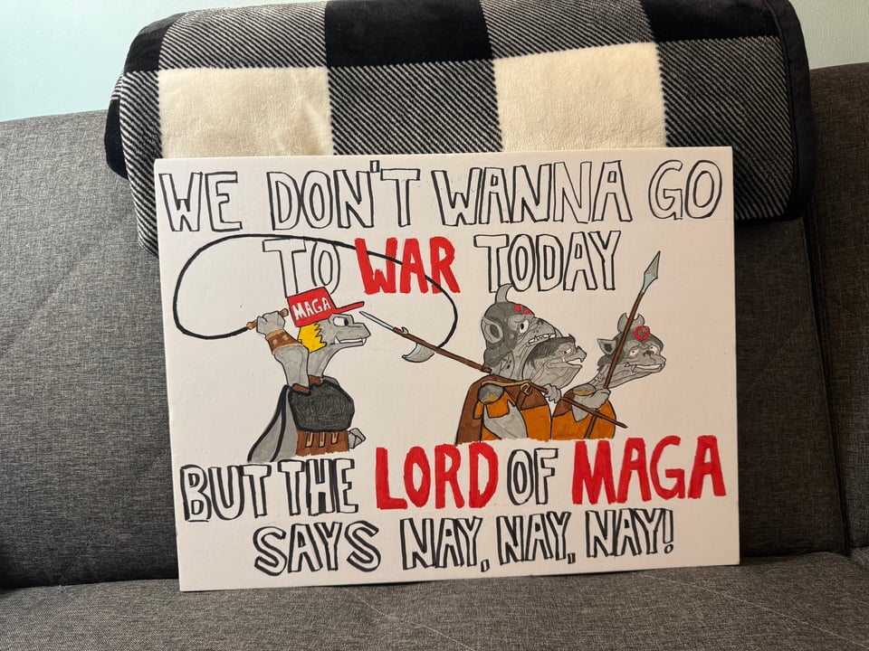 Photo of a protest sign on someone's couch. The sign says "We don't wanna go to war today, but the Lord of MAGA says nay, nay, nay!" It's illustrated with a drawing of the orcs from the animated Return of the King adaptation. The orc wielding the whip is wearing a MAGA hat and has ugly yellow hair.