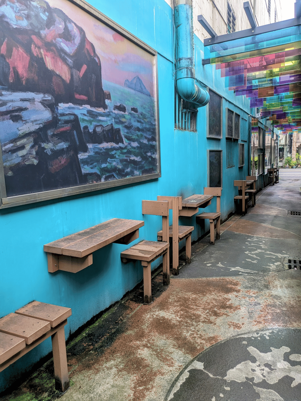 An alley with a bright blue wall and concrete floor. The wall of the alley has half of tables and chairs sticking out of it as if they're stuck in the wall. There's a painting of cliffs by an ocean on the wall. In the background, colored squares of plastic hang rainbow like above the alley.