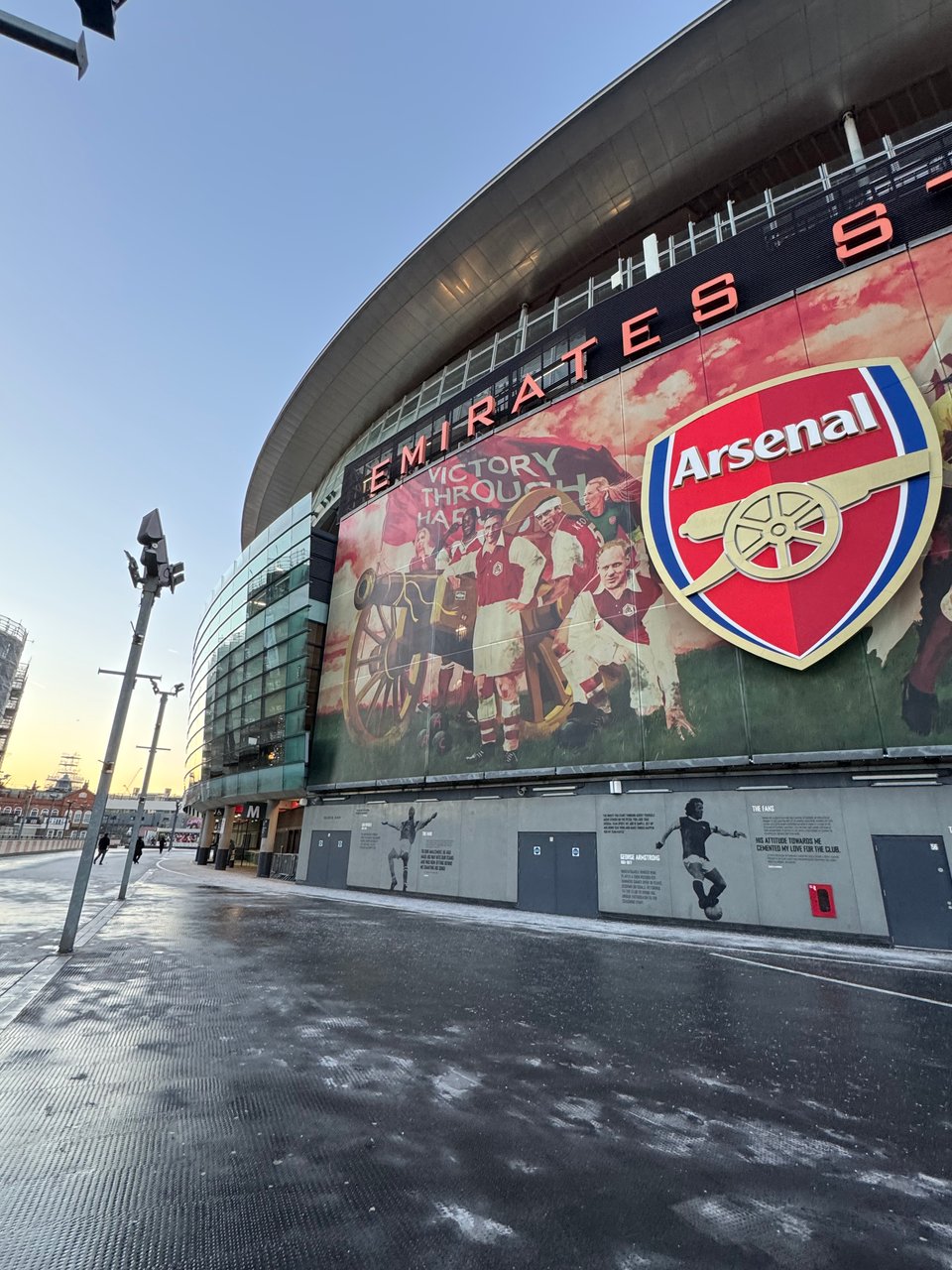 External shot of Arsenal’s Emirates Stadium. An impressive stand with a giant red Arsenal crest sits in front of a clear blue winter sky.
