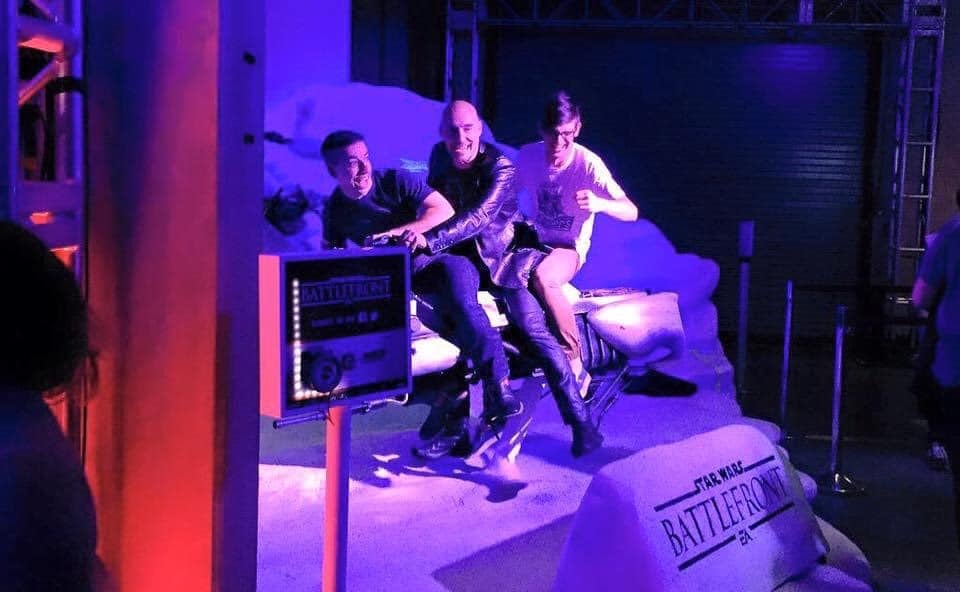 Grant Morrison and two pals laughing on a speeder bike replica at a party, with purple and red lighting, and the logo Star Wars Batllefront