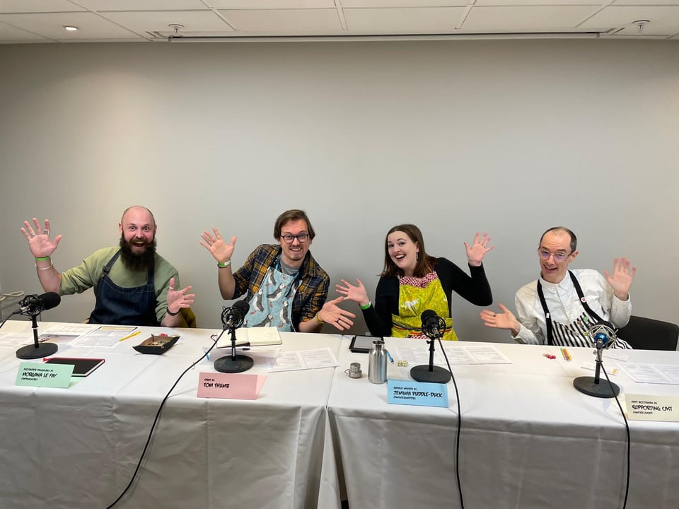 Alex, Strat, Nat and Matt sat behind a recording table, wearing aprons and doing jazz hands