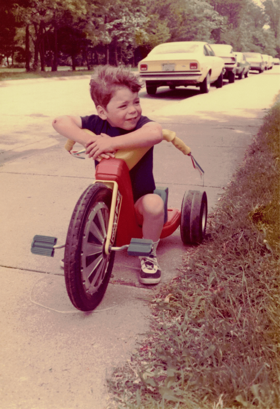 An archival photo from the 1970s of a young blonde boy of perhaps four or five years old sitting on a Big wheel, A plastic tricycle with a big thick front wheel, a red body, Two smaller black wheels, and bright yellow handlebars with trailers sticking out from the edges of them.
