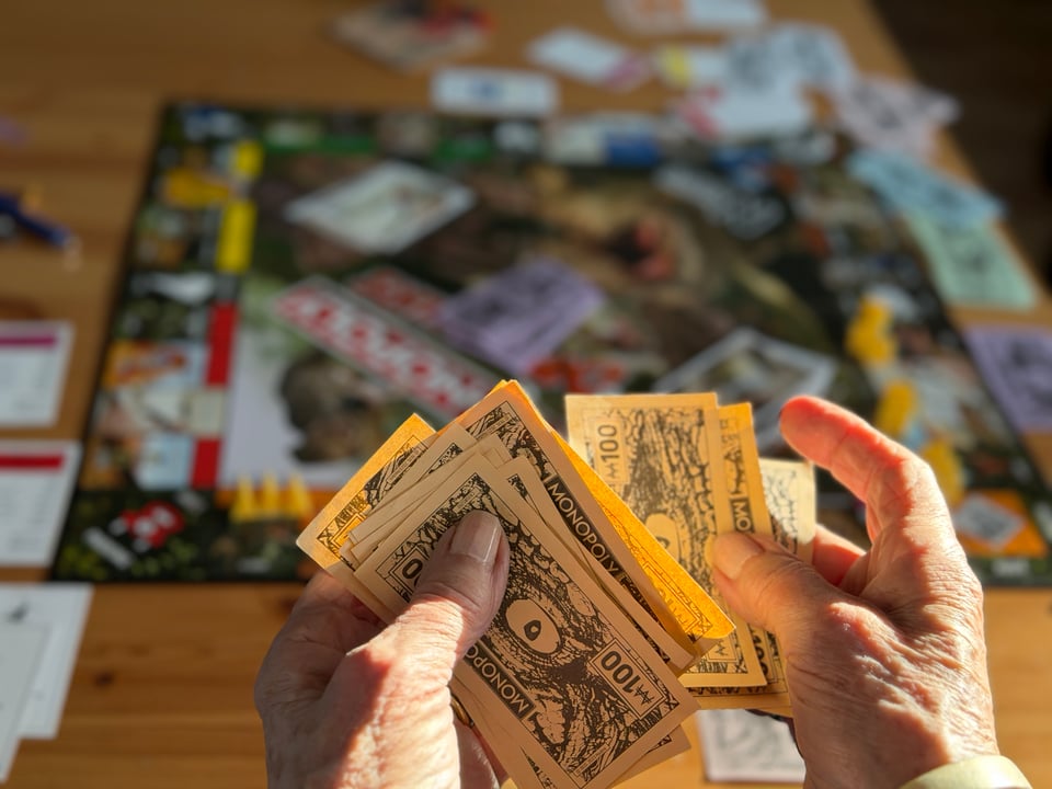hands hold a large pile of "100" Monopoly bucks and the monopoly board is blurred in the background.