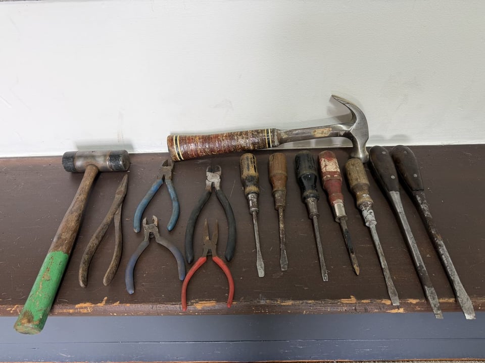 Fourteen salvaged hand tools laid out on a porch rail - two hammers, five pliers and cutters, and seven screwdrivers