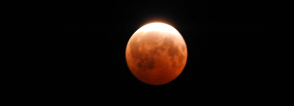 A close up photo of a partial lunar eclipse, with the moon shaded in rusty red and a brighter arc at the top, showing where the earth's shadow doesn't quite cover it.