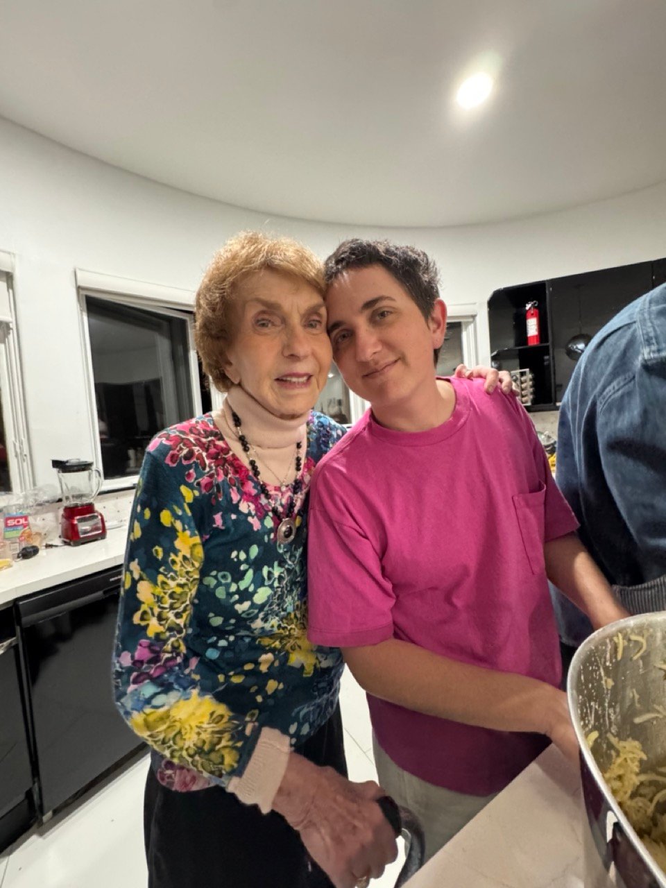 an older white woman in a flowery top holding a cane smiling at the camera. Her arm is around a 30 something white person in a bright pink top, also smiling at the camerar