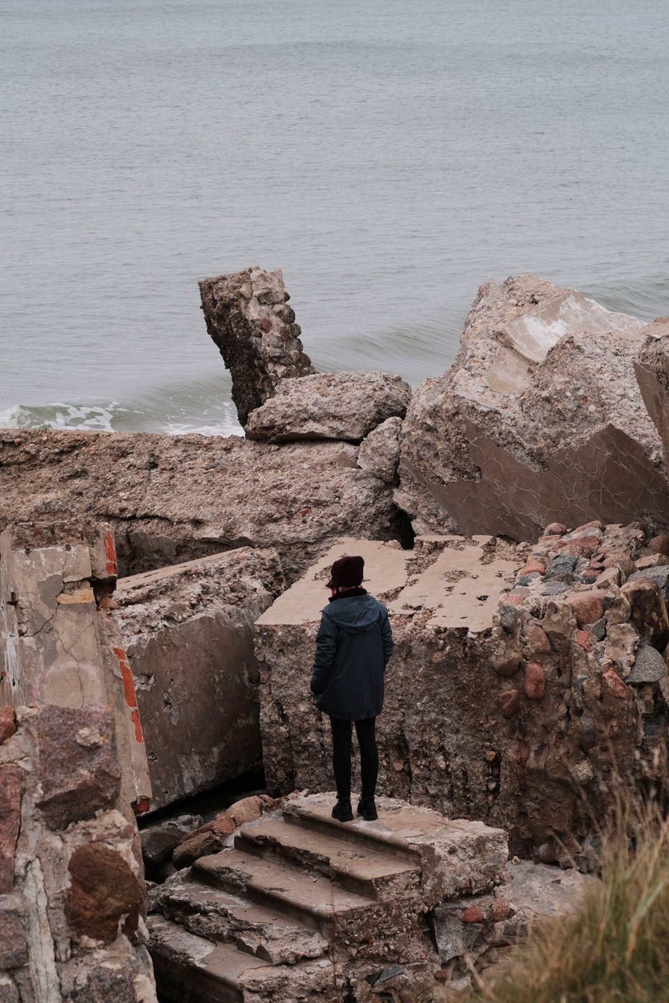 A photo of a person standing on the disconnected remains of stairs, among tall stacks of concrete rubble, with the sea looming behind it all.