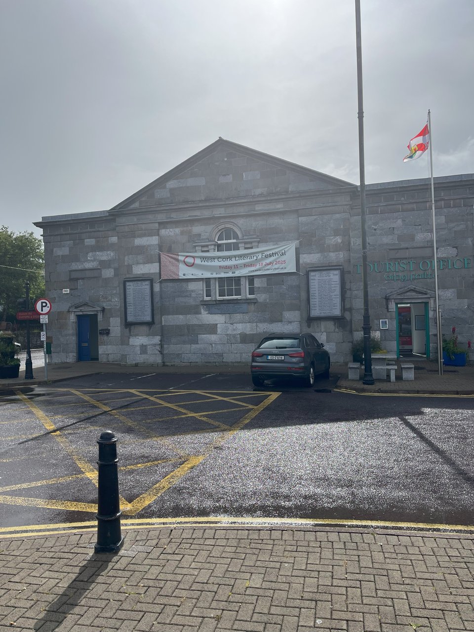 A photograph of the Tourist Office in Bantry. It is made of old grey stone. There is a glare from the sun breaking through the overcast sky, and reflecting on the wet tarmac. On the building, a white banner reads, "West Cork Literary Festival".