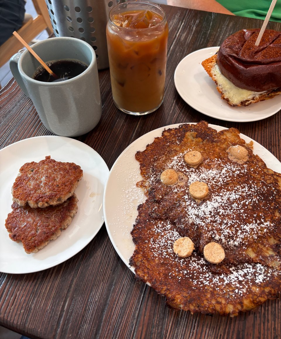 array of breakfast dishes — sausage patties, oat cakes with pats of maple butter, hot coffee, iced coffee, and an egg sandwich — on wooden table top