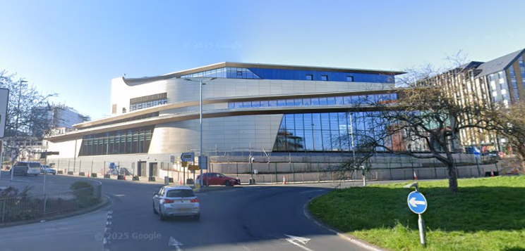 A large building overlooking a roundabout. Each level slopes in different ways, and the walls curve around corners. Some of the walls are curtain glass, others are clad in a pale bronze coloured metal. It glows like a starship.