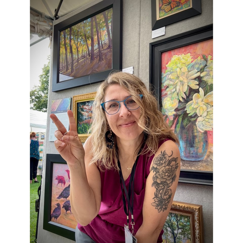 Mary Pow in her art fair booth making a peace sign and smiling at the camera