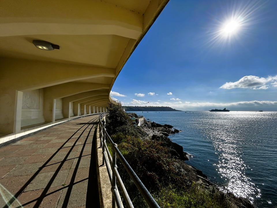 A very sunny day, looking out towards Plymouth Sound. To one side, cream cantilevered concrete creates a colonnade over yellow and red chequered paving.