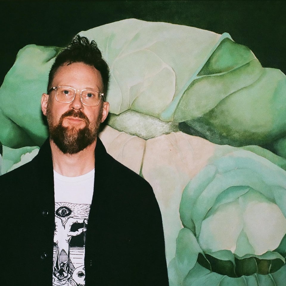 A man with short hair, a beard and glasses stands in front of a backdrop featuring shades of pale green.