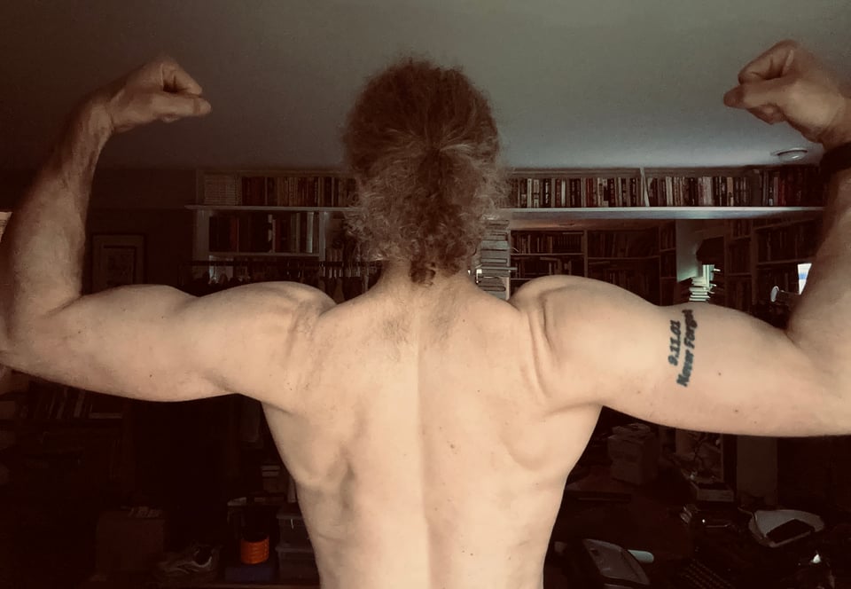 Photo of white man’s back and arms while he’s flexing. He’s got long curly hair tied back in a ponytail, and his shoulders look pretty good