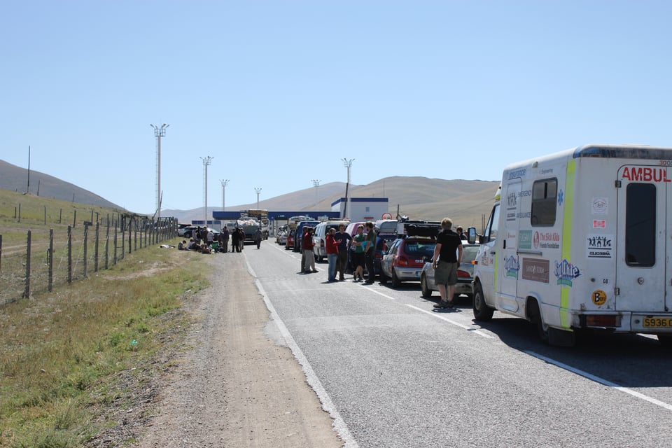 Photograph of a long queue of vehicles. In the mid distance are the buildings, fences, and floodlights of a border checkpoint. In the far distance are some rolling green hills.
