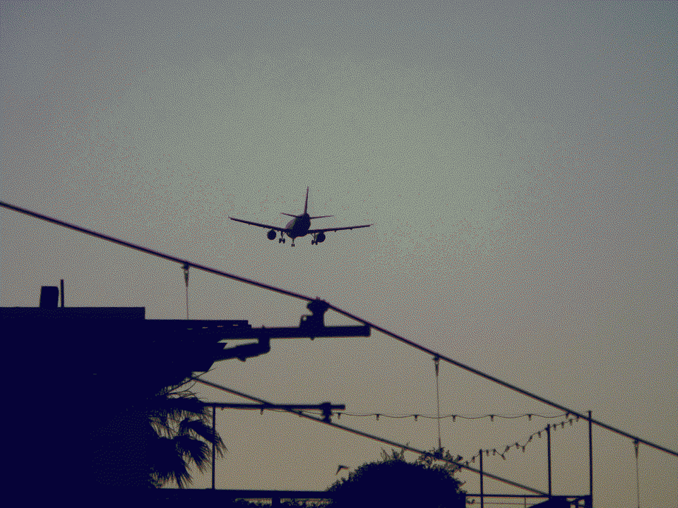 The silhoutte of a commercial jet descending at dusk. Telephone lines, strings of lights, palm trees, and buildings in the foreground give the impression that the plane is getting very close to the ground.