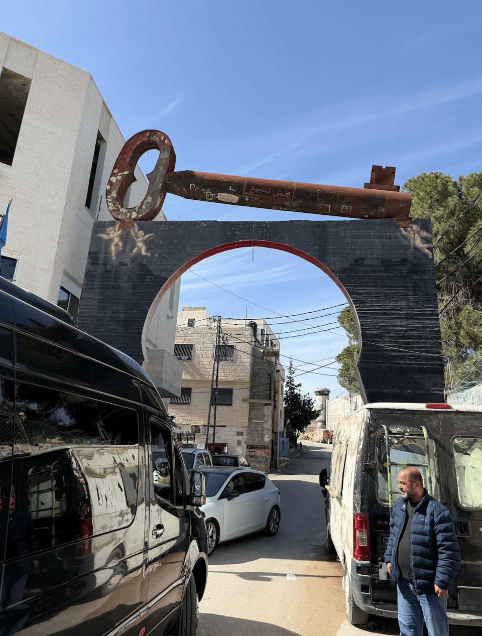 Photograph of a street scene featuring a large black archway with a rusted key sculpture mounted on top. Vehicles and a man in a blue jacket are visible beneath the arch, with buildings and power lines in the background under a clear blue sky.