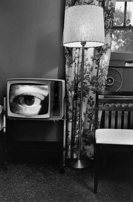 black and white photograph of a room with a television, on the screen is an extremely close image of a man's eye.
