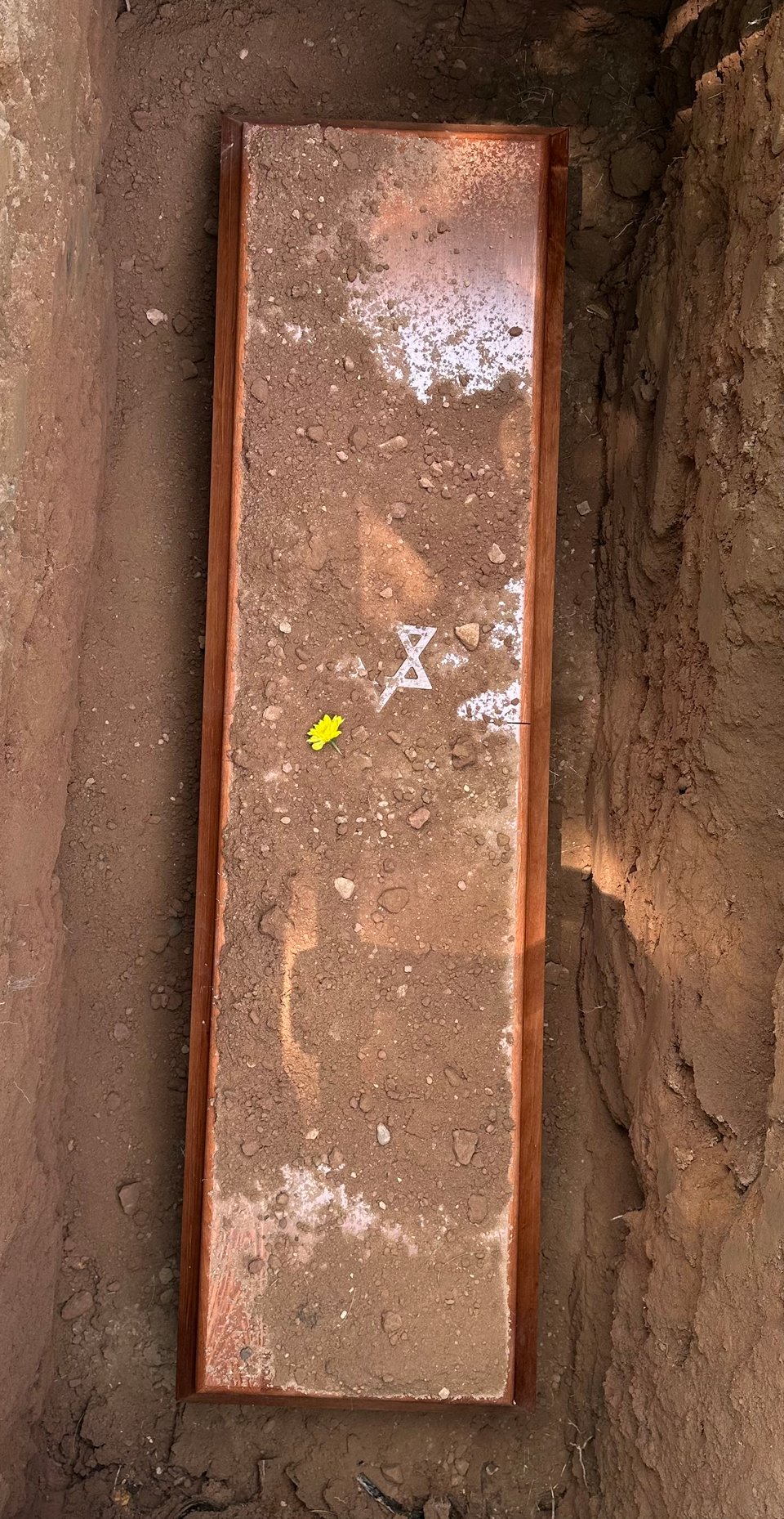 Photo of a simple coffin with a Star of David, in a grave. It’s covered in dirt and a yellow flower lies on top.