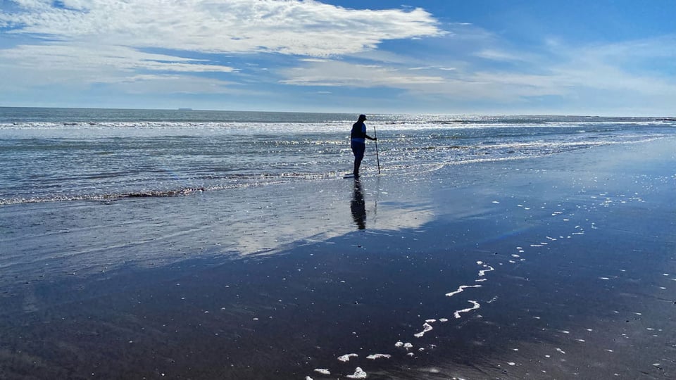 A woman using a walking stick, walks on the beach in the low tide. The blue sky is reflected in the wet sand. The woman is in silhouette.