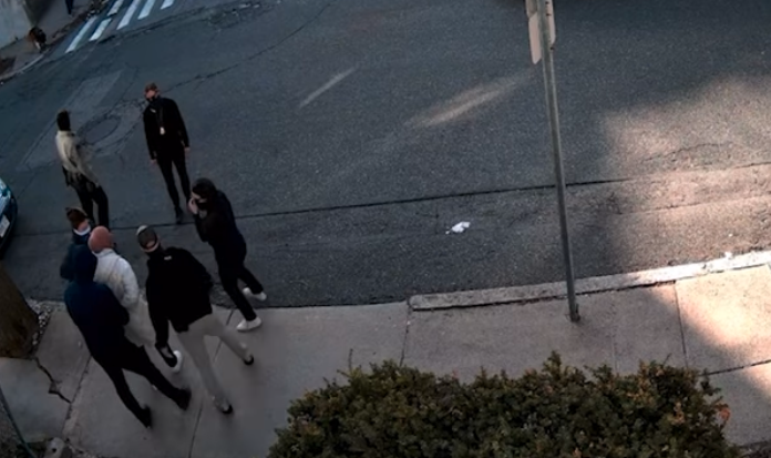 Rumeysa Ozturk, in white jacket and pink head scarf, stands on the sidewalk outside her residence while surrounded by five masked agents. One is putting handcuffs on her.