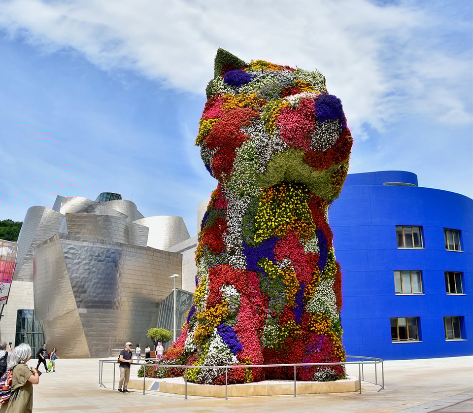 A puppy made of blooming flowers outside of a blue-and-titanium museum.