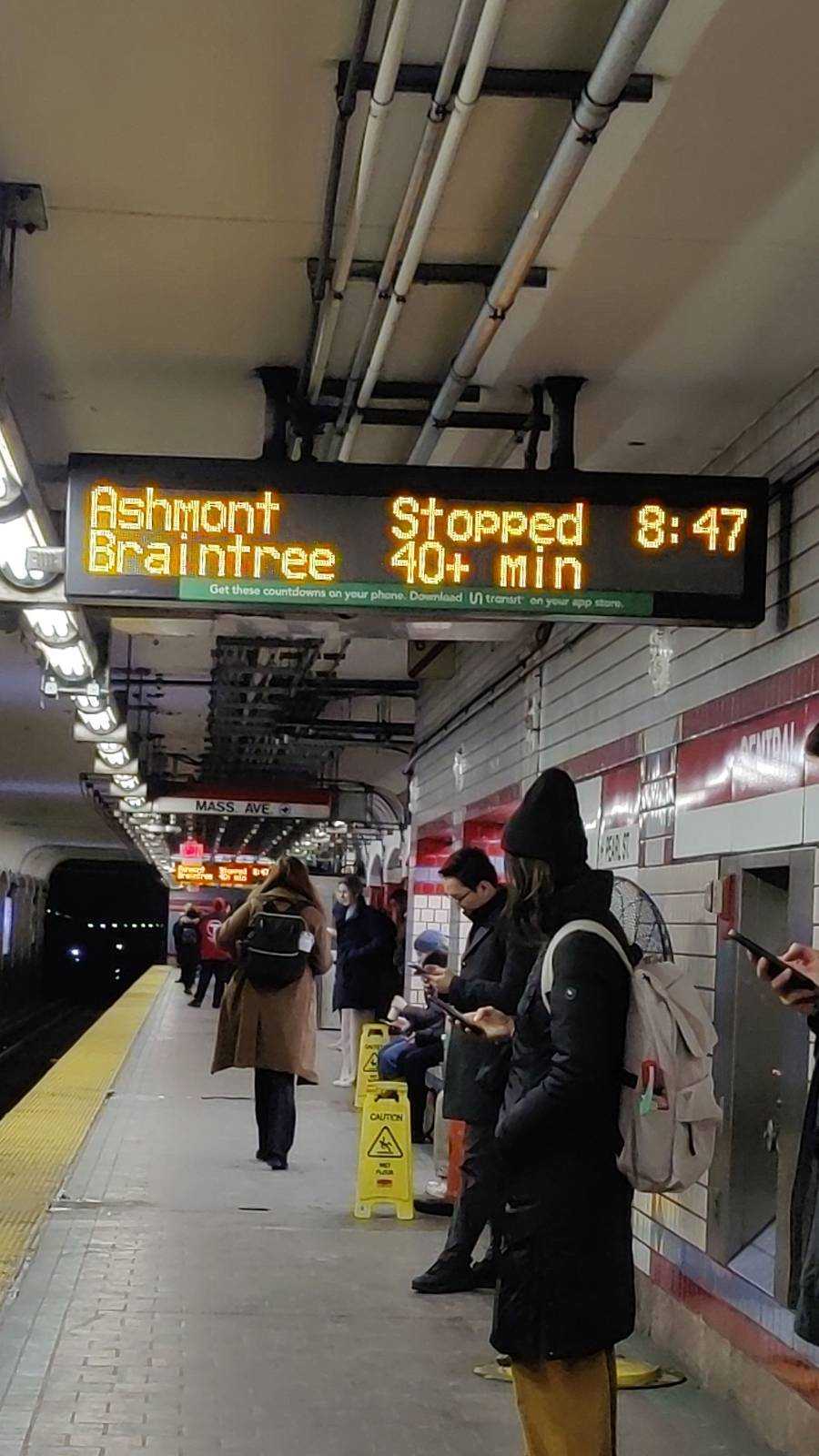 Several commuters, bundled in their winter coats, stand under a sign telling them their train is stopped and it will be 40+ minutes until the next train.