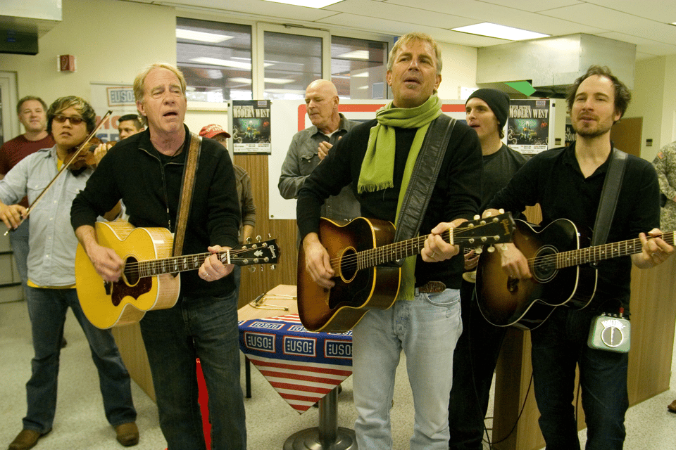 Kevin Costner and his band. Kevin and two other men have guitars, and there's a fiddle player in the background. They appear to be in some sort of municipal building with cheap shelves in the background and a suspended ceiling built out of cheap foam panels. The carpet is the rough, short-pile, grey kind that is only ever used for meeting rooms or similar.