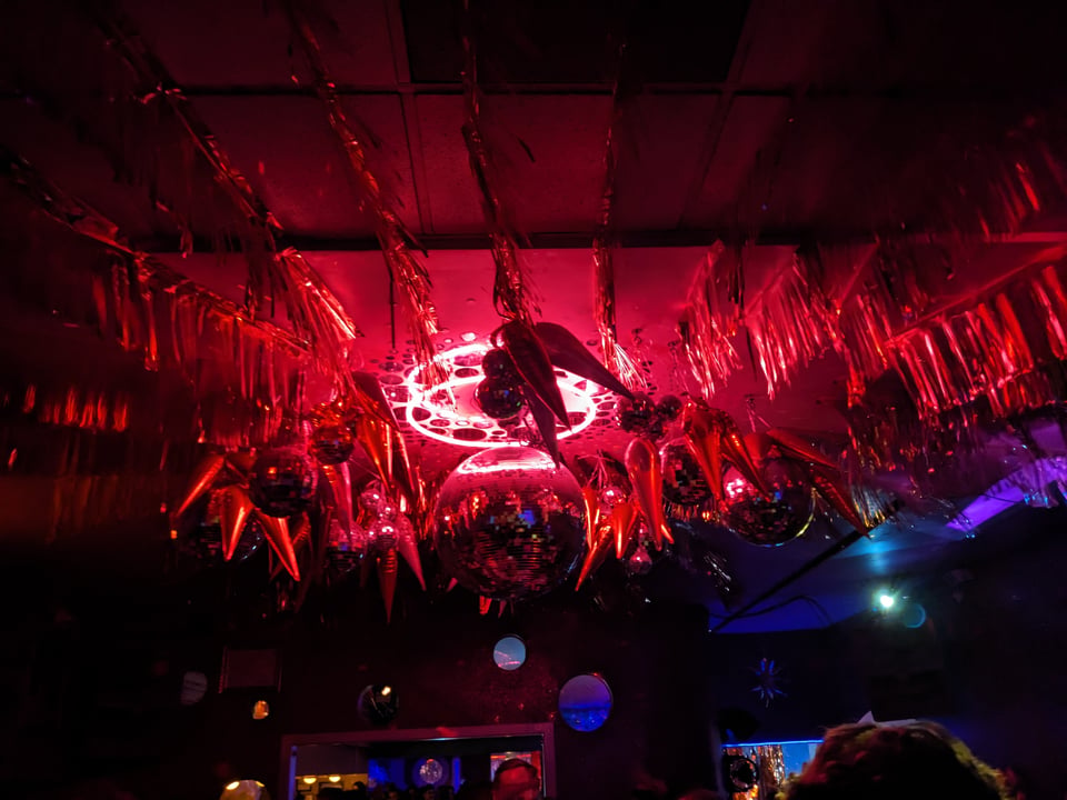 a colorfully lit yet also dark view of the ceiling of a bar, with a disco ball, some balloons and streamers, and mostly red neon lighting with some purple and blue in the mix, plus a few people's heads.
